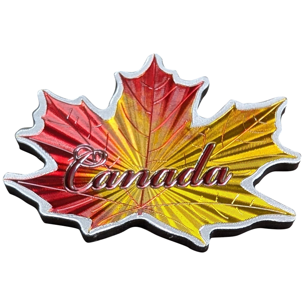 A maple leaf-shaped Canada souvenir magnet or pin with a metallic red, yellow, and orange gradient, featuring the word 'Canada' in metallic script across the center, set against a plain white background.